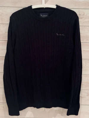 Svart kabelstickad tröja Bondelid - Kabelstickad svart tröja från Bondelid med rund halsringning och diskret broderad logga på bröstet|||Tröjan har långa ärmar och ribbade muddar vid ärmslut och nederkant|||Perfekt för dig som gillar klassisk och stilren design||| Hör av dig vid minsta lilla fråga /// GC