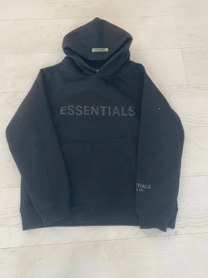 Svart Essentials Fear of God hoodie - Svart hoodie från Essentials Fear of God med stor logga på bröstet och mindre tryck på ärmen. Klassisk huva och känguruficka framtill. Tillverkad i mjukt bomullsmaterial som känns skönt mot huden. Perfekt för en avslappnad och trendig streetwear-look.