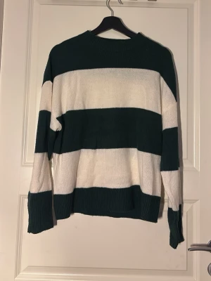 Randig stickad tröja i grönt och vitt - Stickad tröja med breda horisontella ränder i mörkgrönt och vitt. Tröjan har rund halsringning, långa ärmar och ribbstickade muddar. Perfekt för en avslappnad och trendig look.