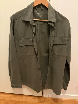 Grön linneskjorta från A Day's March - Säljer en grön skjorta i linne från A Day's March. Skjortan har två bröstfickor med lock, klassisk krage och knäppning framtill. Perfekt för dig som gillar en avslappnad men stilren look.