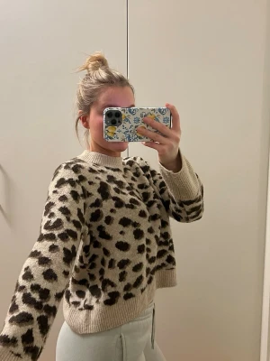 Beige leopardmönstrad stickad tröja - Snygg stickad tröja i beige med leopardmönster i svart och brunt. Tröjan har rund halsringning, långa ärmar och ribbade muddar vid ärmslut och nederkant. Perfekt för dig som gillar djurmönster och vill ha en trendig look.