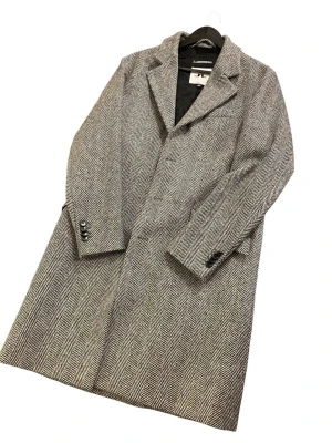 Grå ullrock från J.Lindeberg  - Snygg grå rock från J.Lindeberg i klassiskt fiskbensmönster. Rocken har tre knappar framtill, breda slag och två fickor med lock. Tillverkad i ullblandning och helfodrad för extra komfort. Perfekt för kyliga dagar och stilrena outfits. Modellen är 181