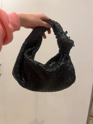 Svart flätad handväska - Trendig svart handväska med flätad design och rundad form. Väskan har ett kort handtag och är tillverkad i ett material som liknar läder. Perfekt för dig som gillar en stilren och modern look.