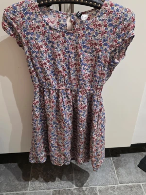 Blommig kortklänning i bomull - Söt kortklänning med blommigt mönster i rött, blått och lila på ljus botten. Klänningen har rund hals, korta ärmar och markerad midja. Tillverkad i mjuk bomull, perfekt för varma dagar. Enkel att styla med sneakers eller sandaler.