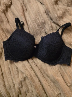 Svart spets-bh med vadderade kupor - Snygg svart bh med vadderade kupor och vacker spets över hela ytan. Justerbara axelband och liten dekorativ detalj mellan kuporna. Perfekt för dig som gillar en feminin och stilren look. Använd Max 3 ggr