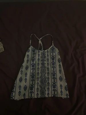 Vit och blå mönstrad linnetopp - Snygg vit linnetopp med blått etno-inspirerat mönster och smala axelband som korsas i ryggen. Toppen har en luftig passform och är perfekt för varma dagar. Materialet känns lätt och bekvämt mot huden.