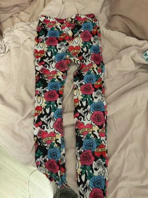 Färgglada leggings med rosor och text - Säljer ett par leggings med färgglatt mönster av röda och blå rosor, hjärtan och banderoller med texten 'Love Me'. Basen är svartvit med inslag av grönt och gult. Tight passform och stretchigt material, perfekta för dig som gillar att sticka ut.