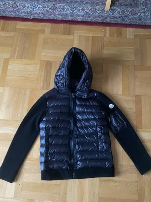 Svart dunjacka med huva från Moncler - Snygg svart dunjacka från Moncler med glansig, quiltad framsida och huva. Ärmar och rygg är i mjukt stickat material, vilket ger en cool kontrast. Jackan har två dragkedjefickor fram och en Moncler-logga på ärmen. Perfekt för kalla vinterdagar.