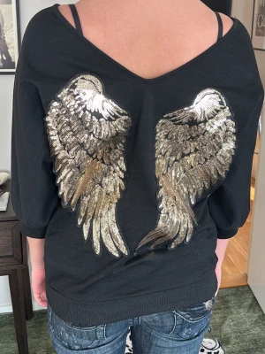 Svart tröja med paljettvingar - Säljer en svart tröja med avslappnad passform och bred urringning som kan bäras off-shoulder. På ryggen finns stora, glittriga paljettvingar i guld och silver. Tröjan har trekvartsärmar och ribbade muddar. Perfekt för den perfekta 2000s looken💕💕
