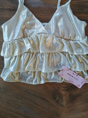 Glittrig guldig linnetopp från SHEIN - Supersnygg glittrig linnetopp i guld från SHEIN med volanger och tunna axelband. Toppen har en v-ringad skärning. Materialet är skimrande och känns lätt på kroppen. Helt ny, aldrig använd.