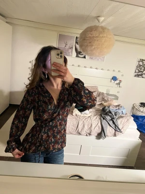 Blommig omlottblus  - Snygg omlottblust med blommigt mönster i vinrött, rosa, blått och gult på mörk botten. Blusen har långa ärmar och knytband i midjan för en figurnära passform.