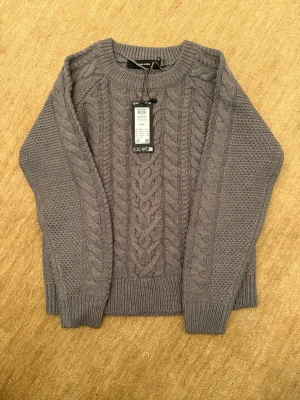 Grå kabelstickad tröja Vero Moda - Säljer en grå kabelstickad tröja från Vero Moda med grov struktur och rund halsringning. Tröjan har breda muddar vid ärmslut och nederkant samt ett klassiskt flätmönster framtill. Perfekt för lager på lager och riktigt mysig.