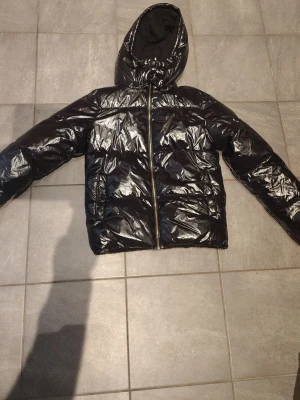 Svart glansig pufferjacka med huva - Säljer en svart pufferjacka med glansig finish och huva. Jackan har dragkedja framtill och två sidofickor med tryckknappar. Perfekt för kalla dagar och ger en cool streetwear-look. Materialet är syntetiskt och jackan är vadderad för extra värme.