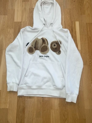 Vit hoodie från Palm Angels med björn - Säljer en vit hoodie från Palm Angels med ett stort broderat björnmotiv på bröstet och texten 'Palm Angels' under. Hoodien har huva med dragsko och en stor magficka. Tillverkad i mjuk bomull, perfekt för en avslappnad streetwear-look.