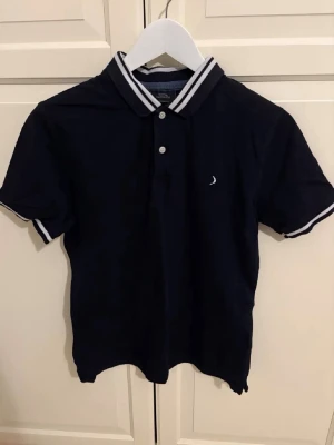 Marinblå pikétröja från bommerang - Klassisk marinblå pikétröja från bommerang med vit logga på bröstet. Tröjan har korta ärmar, vit- och marinrandiga detaljer på krage och ärmslut samt knäppning framtill. Perfekt för en sportig och stilren look.