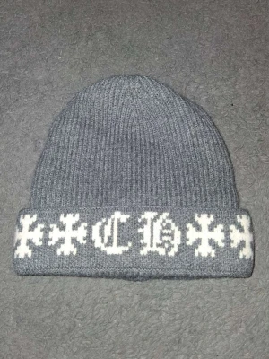 Grå Chrome Hearts mössa med vita gotiska detaljer - Säljer en ribbstickad grå mössa med uppvikt kant och vita gotiska bokstäver samt korsliknande mönster runt om. Perfekt accessoar för dig som gillar streetwear och vill sticka ut lite extra. Materialet känns mjukt och bekvämt.