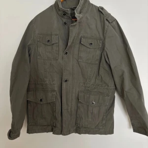 Grön vindjacka med fickor - Snygg grön vindjacka med fyra stora fickor framtill och axelklaffar. Jackan har hög krage, knappar och dragkedja framtill. Perfekt för dig som vill ha en stilren och praktisk jacka till vardags. Längd: 69cm, P2P: 52cm