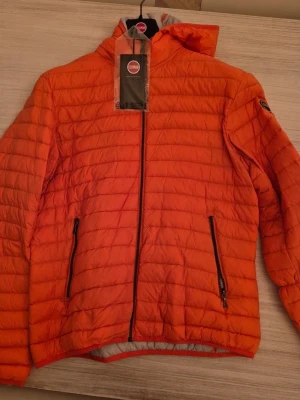 Orange pufferjacka från Colmar - Snygg orange pufferjacka från Colmar med quiltad design och huva. Jackan har dragkedja framtill och två sidofickor med dragkedja. Perfekt för kyliga dagar och ger en sportig vibe. Colmar-logga på ärmen för extra stilpoäng.