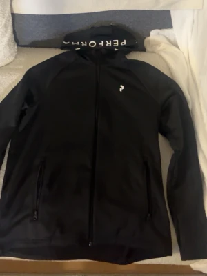 Svart vindjacka från Peak Performance - Snygg svart vindjacka från Peak Performance med dragkedja framtill och två sidofickor med dragkedja. Jackan har en diskret logga på bröstet och huva med tryck. Perfekt för dig som gillar stilren och sportig look.