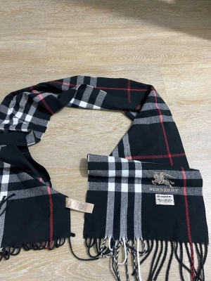  svart rutig halsduk ull - Snygg svart halsduk med klassiskt rutmönster i vitt, grått och rött. Halsduken har fransar i ändarna och är tillverkad i mjuk ull. Perfekt accessoar för att lyfta din outfit med en tidlös och lyxig känsla.