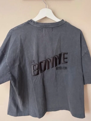  t-shirt från LIS BONNE - Snygg croppad t-shirt i mörkgrå färg från LIS BONNE ATELIER. T-shirten har korta ärmar, rund halsringning och ett stort broderat tryck på ryggen. Tillverkad i mjuk bomull för en avslappnad och trendig look. SLUTSÅLD