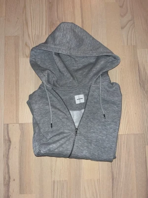 Grå zip-hoodie från Jack & Jones - En stilren grå hoodie med dragkedja från Jack & Jones. Tröjan har huva med dragsko, två fickor framtill och ribbade muddar vid ärmslut och nederkant. Perfekt för en avslappnad look och enkel att matcha med andra plagg.