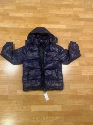 Mörkblå jacka från polo Ralph lauren - Snygg mörkblå dunjacka från polo Ralph lauren med glansig finish och avtagbar huva. Jackan har två stora fickor framtill och dragkedja. Liten röd logga på bröstet. Perfekt för kalla vinterdagar och riktigt varm.