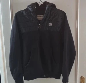 Svart jacka med huva från Moncler - Svart jacka från Moncler med huva och dragkedja framtill. Jackan har quiltade detaljer på axlar och huva samt Moncler-logga på bröstet. Praktiska fickor på sidorna och en sportig look. Perfekt för kyligare dagar.