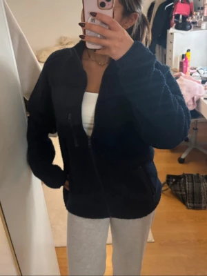 Marinblå teddyjacka med dragkedja - Mysig marinblå teddyjacka med hel dragkedja framtill och hög krage. Jackan har två sidofickor och en bröstficka med dragkedja. Perfekt för kyliga dagar och ger en avslappnad, trendig look. Materialet är fluffigt och mjukt för extra komfort.