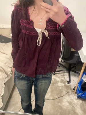 Vinröd blus med broderi och knytband - Supersöt vinröd blus med rosa broderier och dekorativt knytband fram. Blusen har lång ärm och en avslappnad passform. Detaljerad brodyr på axlar och rygg, samt en liten broderad patch bak. Perfekt för en chill och trendig look.