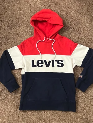 Levi's hoodie i rött, vitt och blått - Snygg hoodie från Levi's med färgblock i rött, vitt och marinblått. Stor logga framtill, huva med dragsko och känguruficka. Perfekt för en avslappnad och trendig stil. Mjuk bomullsblandning som känns skön mot huden. Priset kan diskuteras
