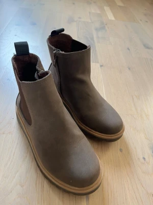Bruna Chelsea boots i mocka - Snygga bruna Chelsea boots med elastiska sidopaneler och dragflik bak. Bootsens ovandel är i mocka och de har en robust sula i gummi. Praktisk dragkedja på insidan för enkel på- och avtagning. Perfekta för dig som vill ha en stilren och enkel look.