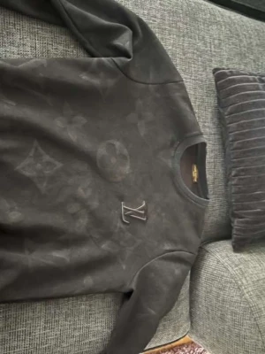 Svart Louis Vuitton sweatshirt - Säljer en svart sweatshirt från Louis Vuitton med diskret monogrammönster över hela tröjan och LV-logga på bröstet. Tröjan har rund halsringning och långa ärmar. Perfekt för dig som gillar exklusiva streetwear-plagg.