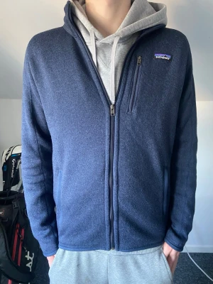 Mörkblå Patagonia fleece  - Mörkblå zip-tröja från Patagonia med bröstficka och diskret logga på vänster sida. Tröjan har hel dragkedja, hög krage och är tillverkad i ett mjukt, stickat material. Perfekt för lager-på-lager och enkel att matcha med andra plagg. Storlek M, passar dig mellan 180-185, mycket fint skick använts fåtal gånger.