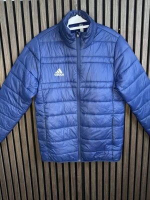 Blå dunjacka från Adidas - Snygg blå dunjacka från Adidas med quiltad design och dragkedja framtill. Jackan har hög krage, två sidofickor med dragkedja och Adidas-logga på bröstet. Perfekt för kyliga dagar och enkel att matcha med olika outfits.