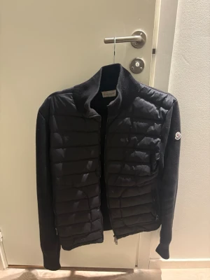 Svart pufferjacka från Moncler - Snygg svart pufferjacka från Moncler med quiltad front och stickade ärmar samt rygg. Jackan har dragkedja framtill, hög krage och Moncler-logga på ena ärmen. Perfekt för kalla dagar och riktigt trendig look. Äkta som alltid!