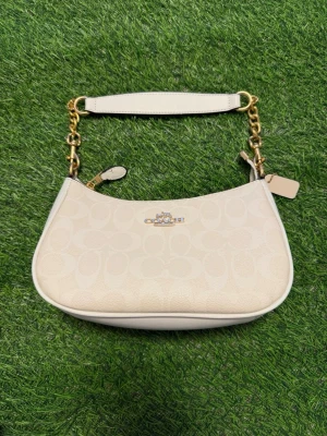 Vit axelremsväska från Coach - Snygg vit handväska från Coach med guldfärgade kedjedetaljer och präglat logomönster. Väskan har en avtagbar axelrem och klassisk Coach-logga framtill. Tillverkad i läder och levereras med original dustbag.