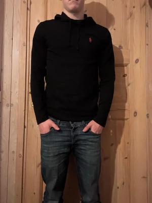 Svart hoodie från Ralph Lauren - Svart hoodie från Ralph Lauren med klassisk huva och dragsko. Liten röd broderad logga på bröstet. Mjuk och skön passform, perfekt till jeans eller joggers. Enkel och stilren design som funkar till det mesta. 