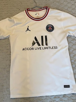 Vit PSG fotbollströja Jordan - Snygg vit PSG fotbollströja från Jordan med svart logga och klubbmärke på bröstet. Röd, vit och svart randig krage. Tillverkad i lätt och ventilerande polyester, perfekt för fotboll eller träning.