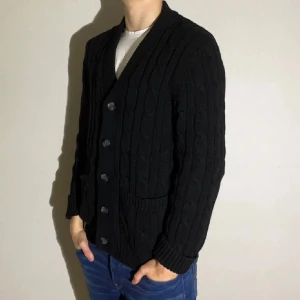 Svart stickad cardigan - Snygg svart stickad kofta med knappar framtill. Den har en klassisk design med ribbade muddar och två fickor. Perfekt för kyligare dagar.