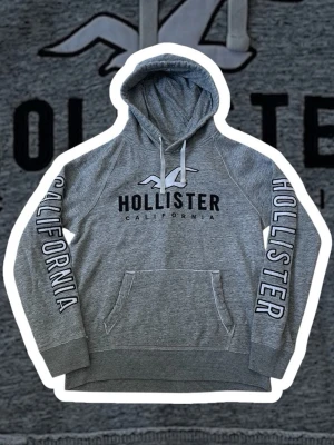 Grå Hollister California Hoodie | L - Snygg grå hoodie från Hollister i storlek L med stor logga och texten CALIFORNIA på bröstet och längs båda ärmarna. Tröjan har huva med snörning och en stor magficka framtill. Mjuk insida och klassisk streetstil.