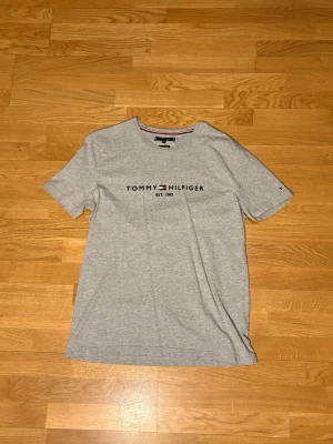 Tommy Hilfiger T‑shirt XXL – sparsamt använd, bra skick - Snygg grå t-shirt från Tommy Hilfiger med klassisk logga och texten 'EST. 1985' broderad på bröstet. T-shirten har rund halsringning och korta ärmar. Perfekt för en clean och stilren look.