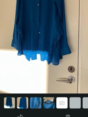 Blå blus med volangdetaljer - Snygg blå blus i ett lätt och luftigt material med knappar framtill och volangdetaljer längs sidorna. Blusen har lång ärm med manschett och klassisk krage. Perfekt för dig som vill ha en stilren men ändå lekfull look.