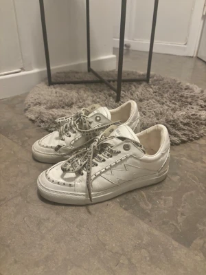 Vita sneakers från Zadig & Voltaire - Snygga vita sneakers från Zadig & Voltaire med rund tå och platt sula. Skorna har coola snören med logotyptext och dekorativa sömmar längs sidorna. Tillverkade i skinn för en exklusiv känsla och passar perfekt till en trendig streetstyle-look.