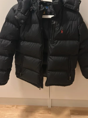 Svart dunjacka från Polo Ralph Lauren - Snygg svart dunjacka från Polo Ralph Lauren med huva och dragkedja framtill. Jackan har det klassiska röda Polo-logot på bröstet och två sidofickor med dragkedja. Perfekt för kalla dagar och riktigt skön att bära.