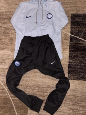 Nike Inter träningsset ljusblå/svart - Säljer ett träningsset från Nike med Inter-logga. Jackan är ljusblå med huva, dragkedja och blå Nike-logga. Byxorna är svarta med resår i midjan och Inter-märke på benet. Perfekt för fotbollsträning eller chill.