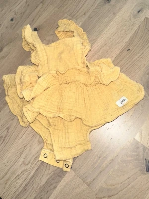 Gul volangbody från Pidde Newbie gul body, storlek 68 - Supersöt gul body med volanger och axelband från Pidde. Tillverkad i mjukt, krinklat bomullstyg och har tryckknappar i grenen för enkel påklädning. Perfekt för dig som gillar lekfulla detaljer och bekväma material.