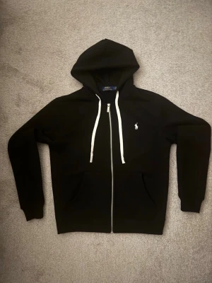 Svart hoodie från Polo Ralph Lauren - Svart hoodie med dragkedja från Polo Ralph Lauren. Klassisk design med vit dragsko i huvan och liten broderad logga på bröstet. Tillverkad i mjukt bomullsmaterial och har två fickor framtill. Perfekt för en avslappnad och stilren look. Small i storleken passar mer som S/M 🇨🇳