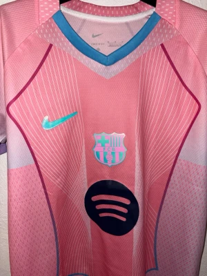 Barça rosa matchtröja Nike Dri-FIT - Snygg rosa FC Barcelona matchtröja från Nike med Dri-FIT-material. Tröjan har turkosa och lila detaljer, klubbmärke och sponsorlogga på bröstet samt blå V-ringad krage. Perfekt för fotbollsträning eller att bära till vardags.