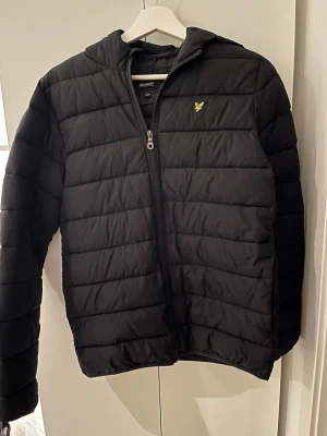 Svart pufferjacka från Lyle & Scott - Snygg svart pufferjacka från Lyle & Scott med gul logga på bröstet. Jackan har huva, dragkedja framtill och quiltad design. Perfekt för kalla dagar och ger en clean streetwear-look. Tillverkad i syntetmaterial för extra värme.